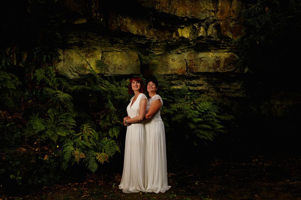 Two brides in white gowns pose back-to-back before a mossy rock wall with lush ferns in a forest setting.