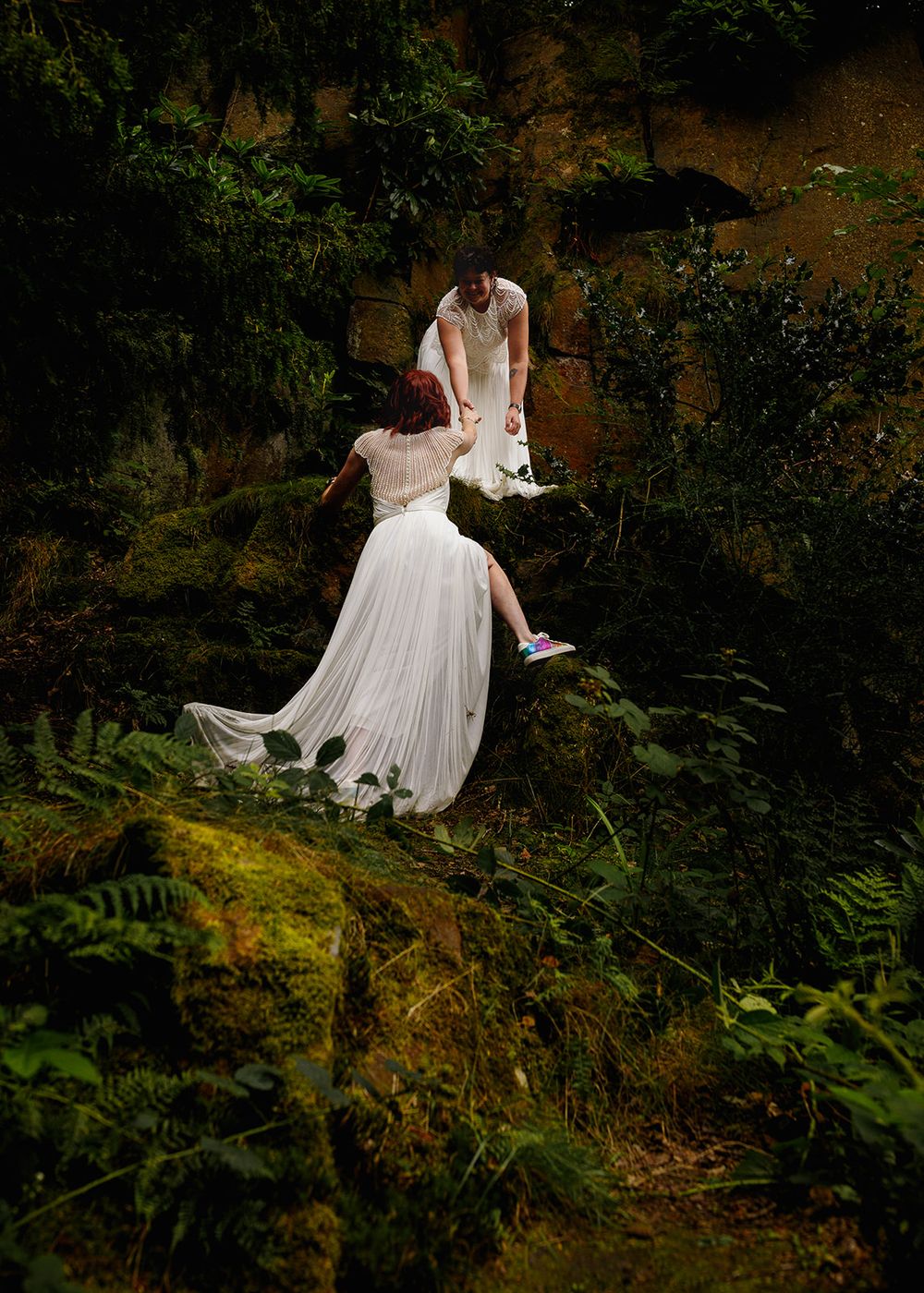 Two brides in white gowns navigate a mossy forest rock, surrounded by lush ferns and greenery.