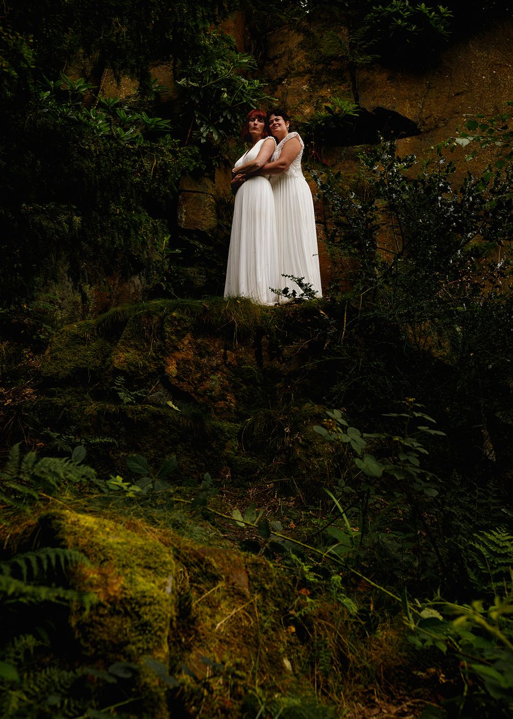 Two brides in white dresses embrace on a mossy rock ledge surrounded by lush green forest foliage.