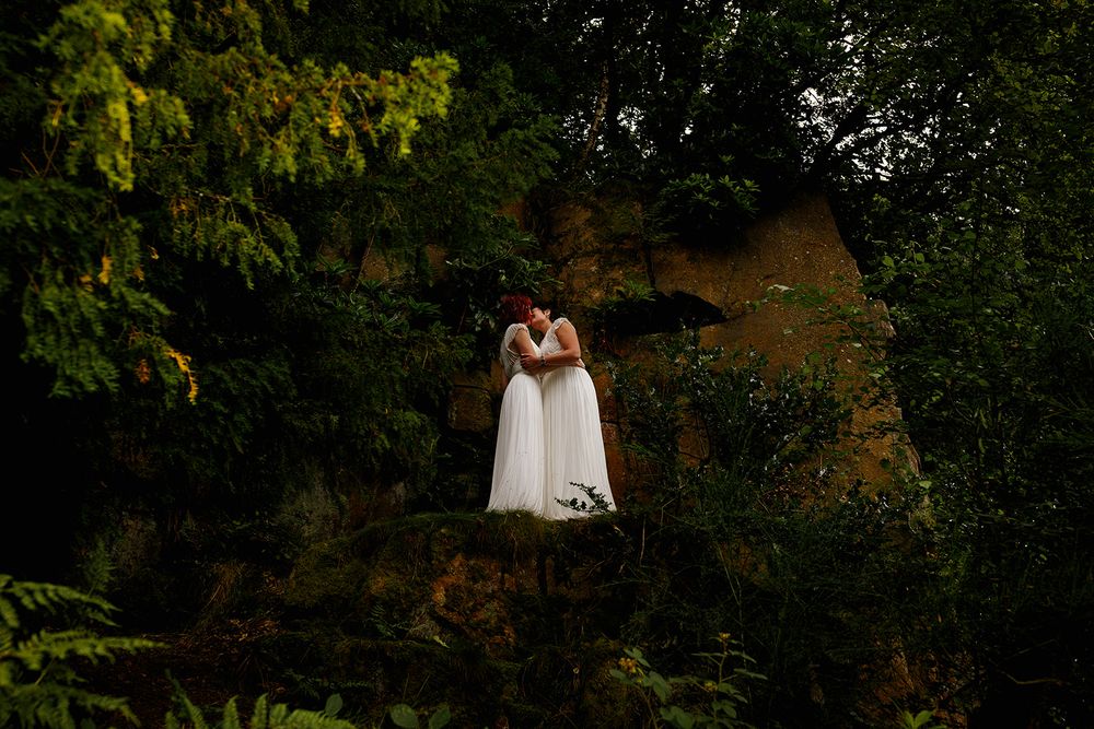Two brides in white dresses share a kiss on a rocky outcrop surrounded by lush green trees.