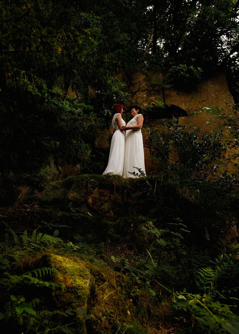 Two brides in white gowns stand together on a mossy rock surrounded by lush green forest and sandstone cliffs.