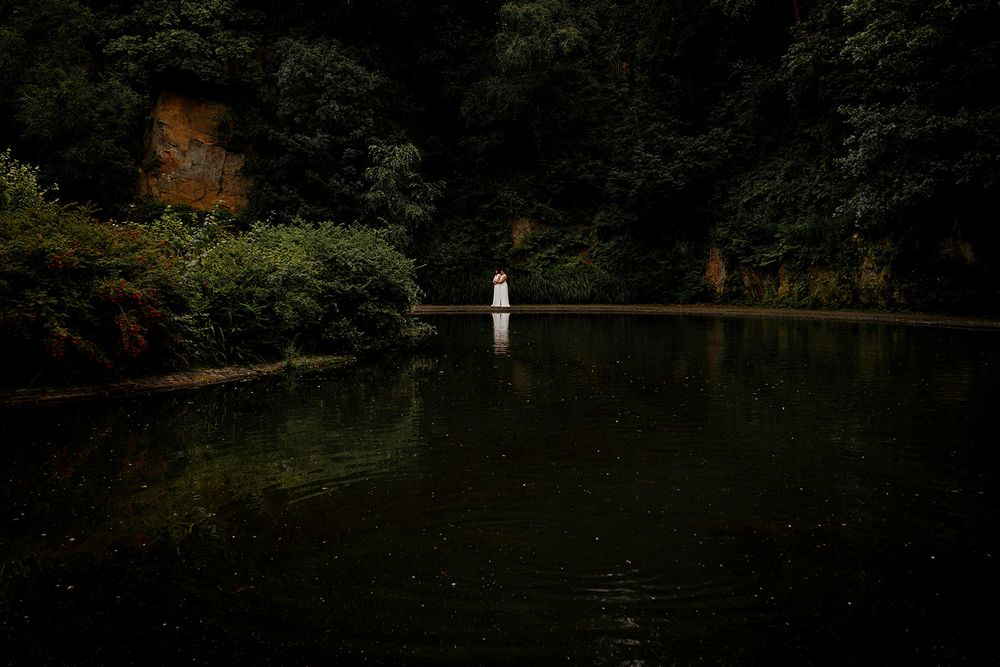 A lone figure in white stands in a dark, reflective lake surrounded by lush green forest and rocky cliffs.