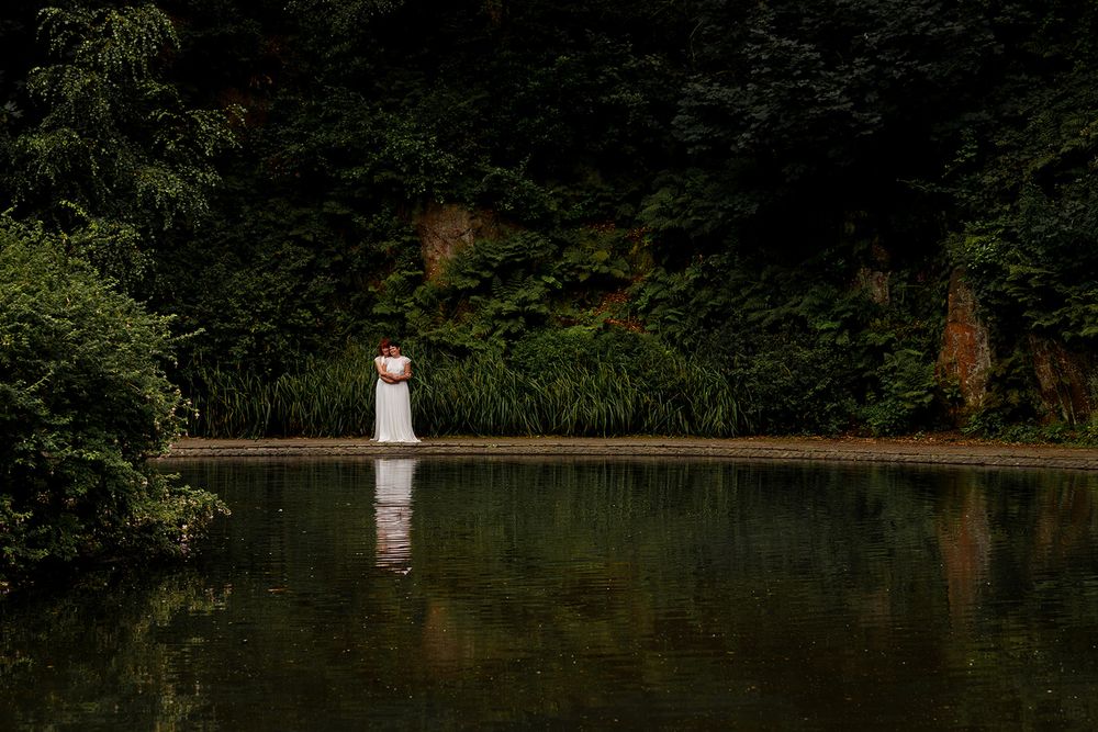 Bride in white dress stands by a reflective pond surrounded by lush green trees in a serene forest setting.
