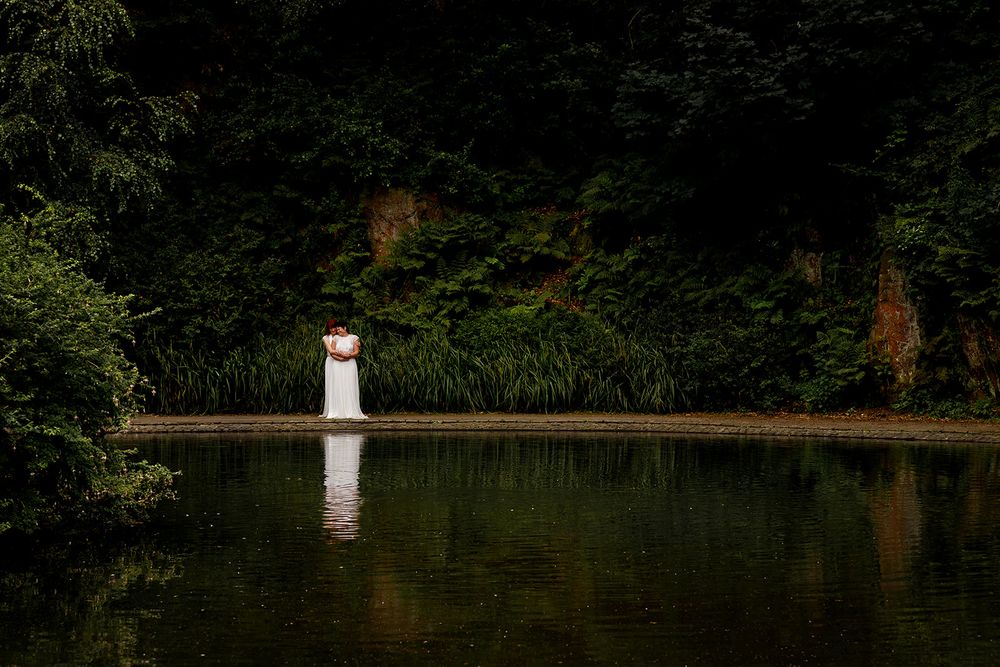 Bride in white dress standing by a serene reflective lake surrounded by lush green trees and foliage.