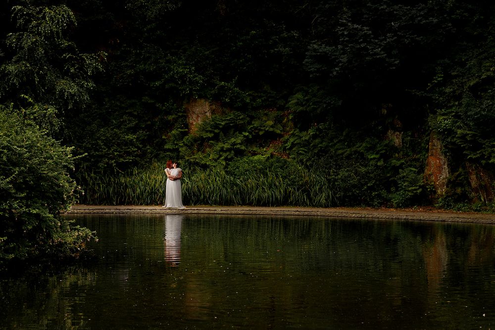 Bride in white gown standing by a tranquil forest pond, surrounded by lush green trees and reflected in the water.