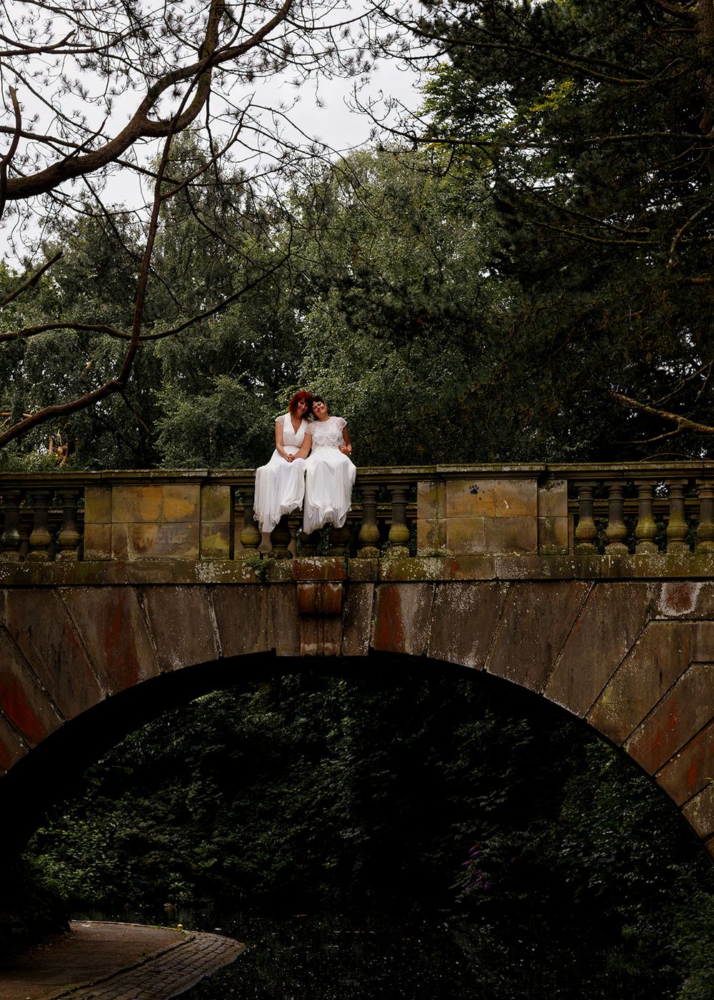 Two brides in white dresses sit together on a stone bridge arch surrounded by lush green trees.
