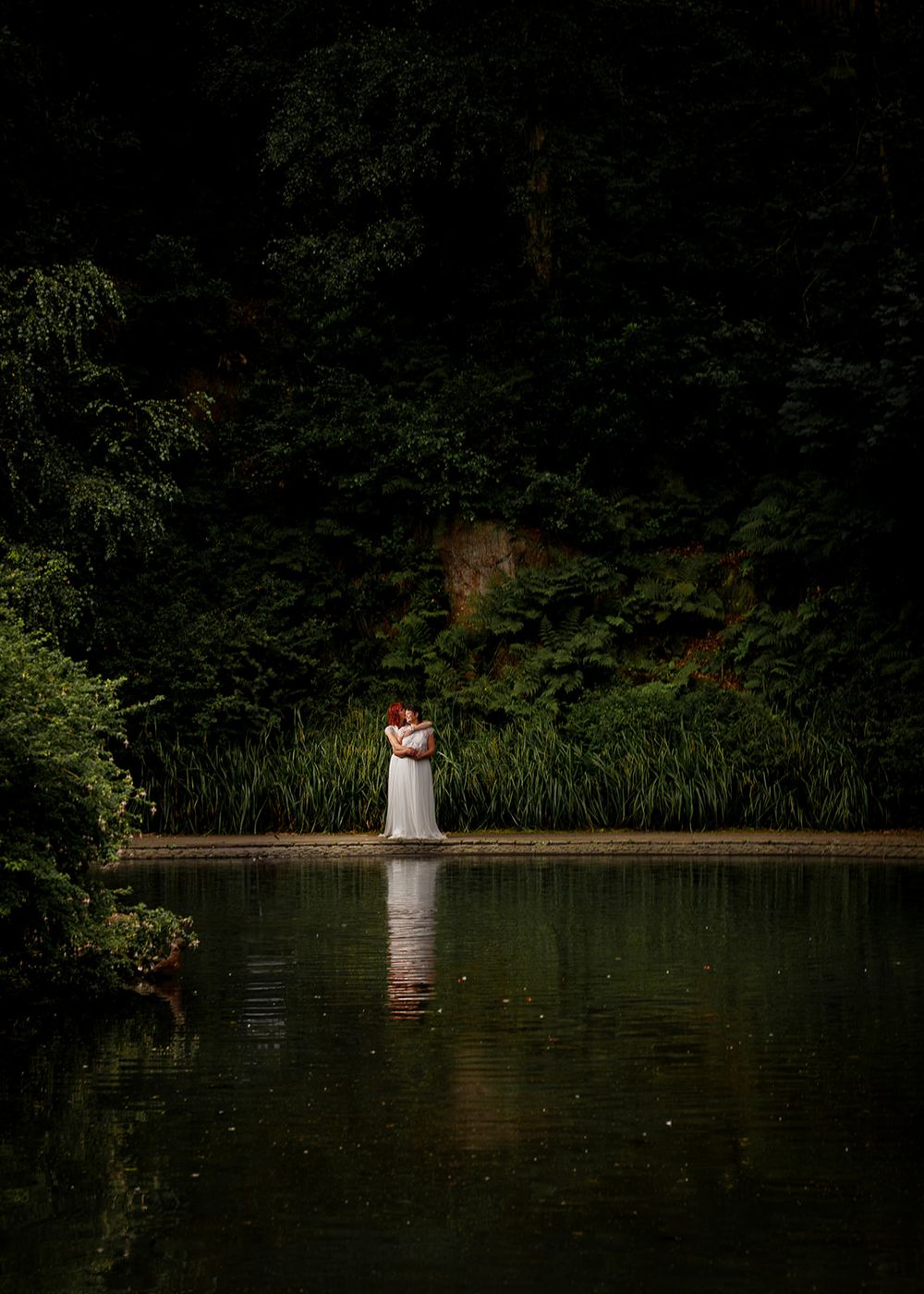 Couple embracing in white wedding attire beside a serene forest pond, surrounded by lush green trees.