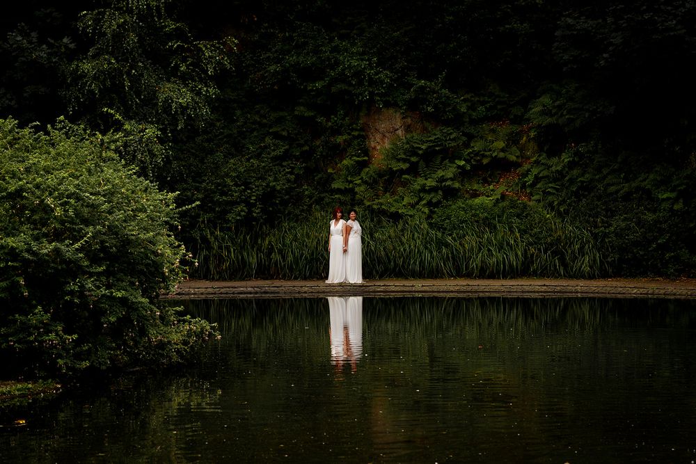 Two brides in white gowns stand by a reflective pond surrounded by lush green forest in a romantic wedding photo.