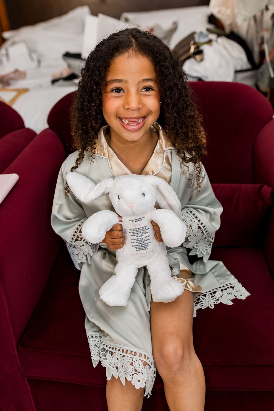 Smiling young girl in sage green lace robe holding white stuffed bunny toy, sitting on red velvet chair.