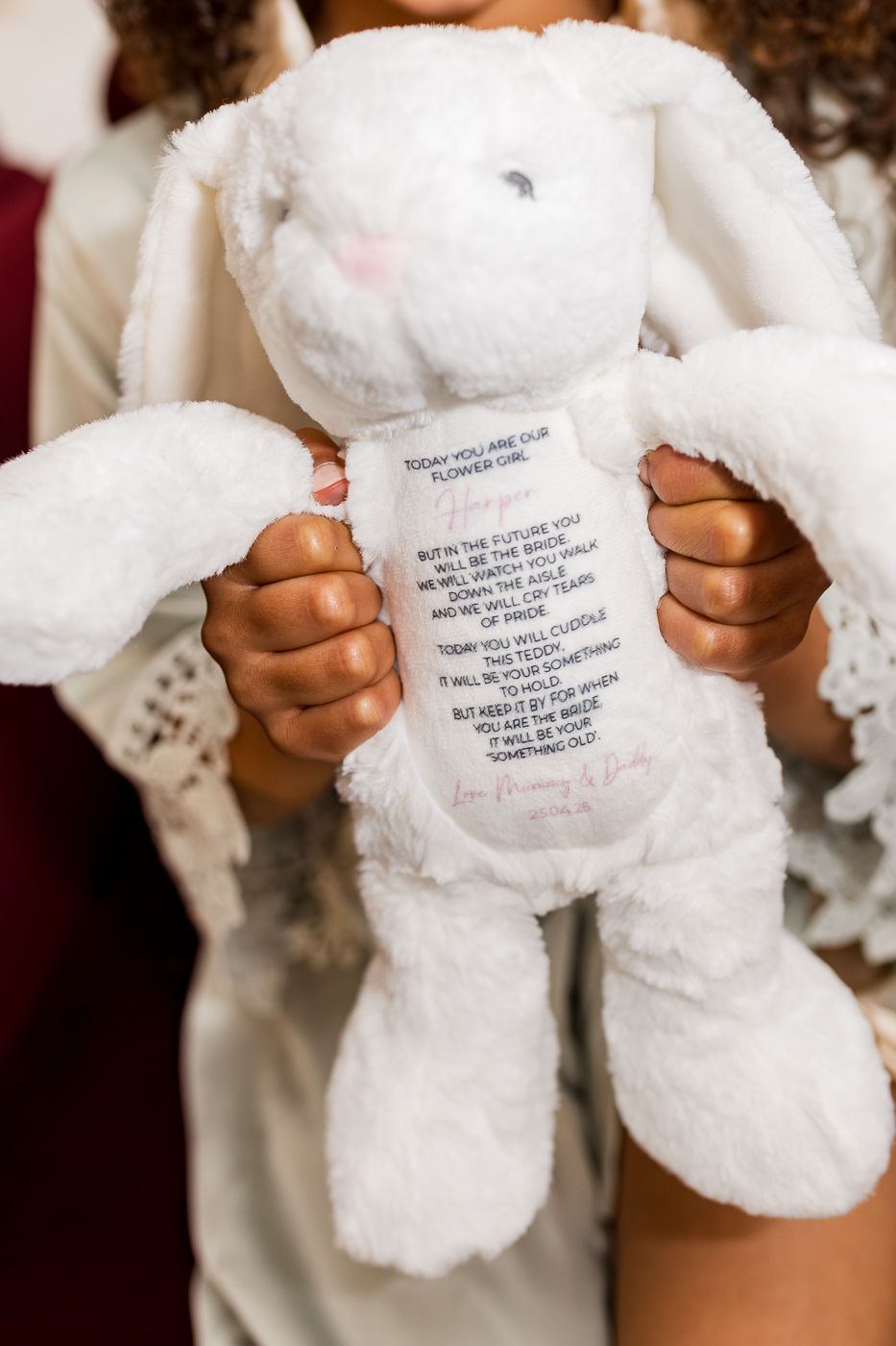 Child holding white stuffed bunny with personalized flower girl message embroidered on its belly at a wedding.