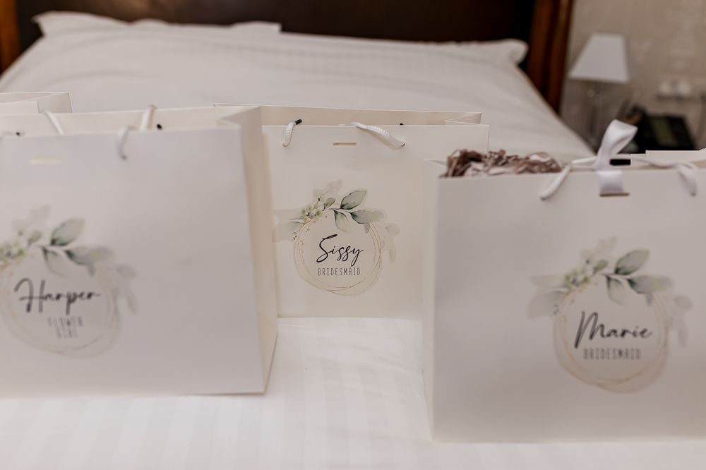 Three personalized white gift bags with floral labels reading Harper, Sissy, and Marie sit on a bed for a bridal party.