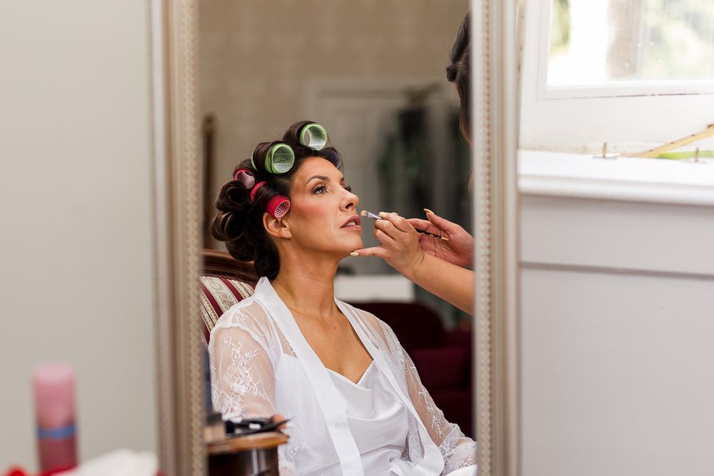 Bride in white robe with pink hair rollers getting makeup applied by a professional artist before her wedding.