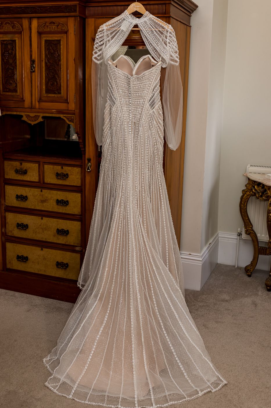 Elegant beaded wedding gown with sheer sleeves and train displayed on hanger against wall near wooden dresser.