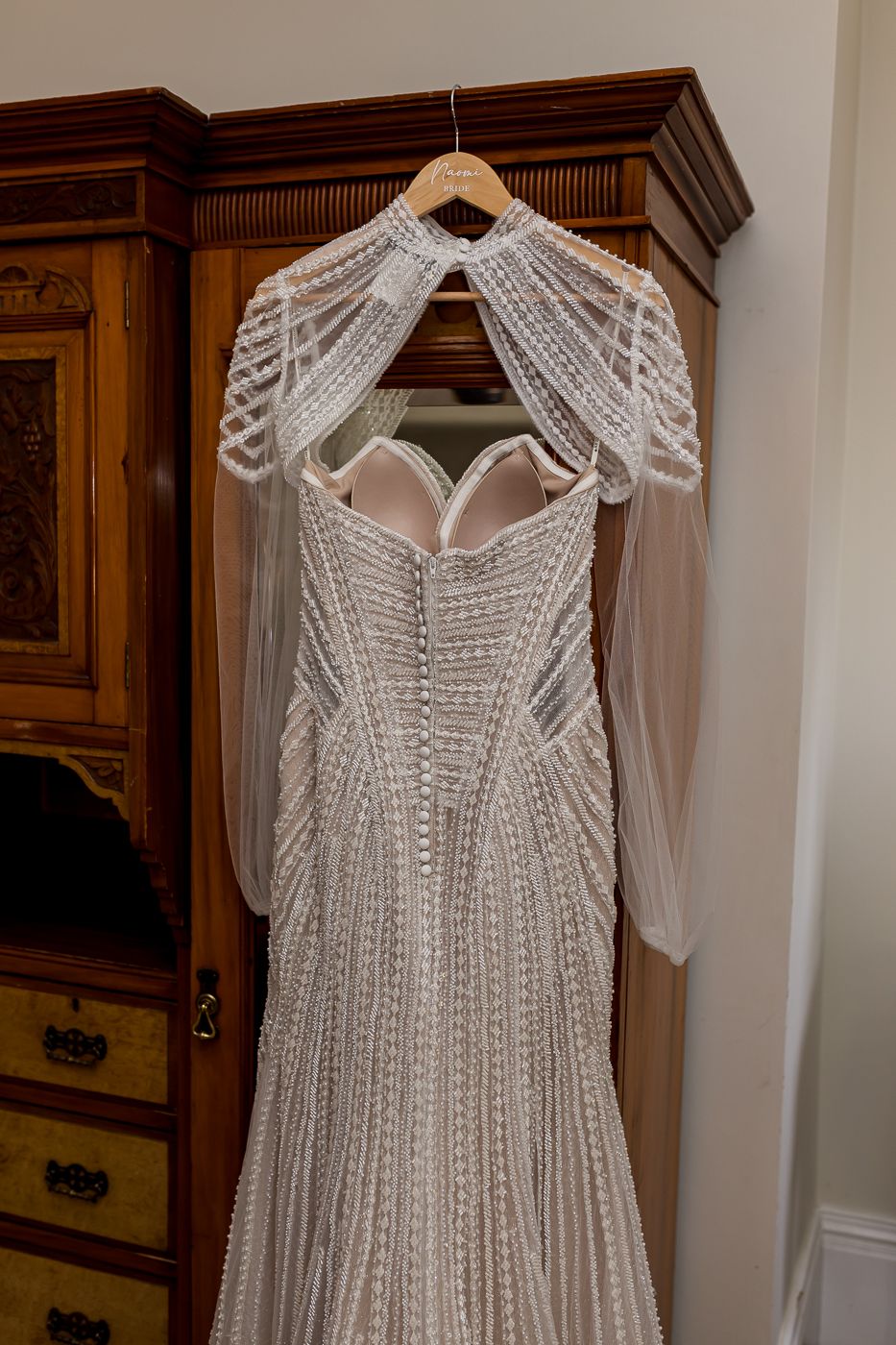 Heavily beaded ivory wedding gown with cape shoulders hanging on wooden armoire hanger.