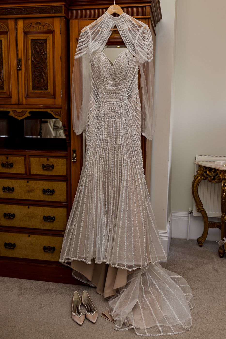 Elegant beaded mermaid wedding dress with high neck and long sleeves hanging on a wooden wardrobe with heels below.