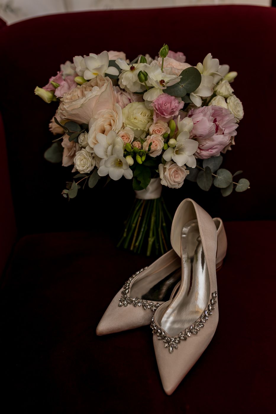 Elegant bridal heels with crystal embellishments beside a romantic pink and white wedding bouquet on a red velvet chair.