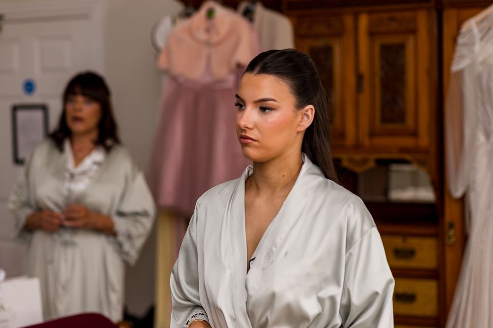 Woman in silver satin robe standing in a bridal dressing room with another woman in pajamas in the background.