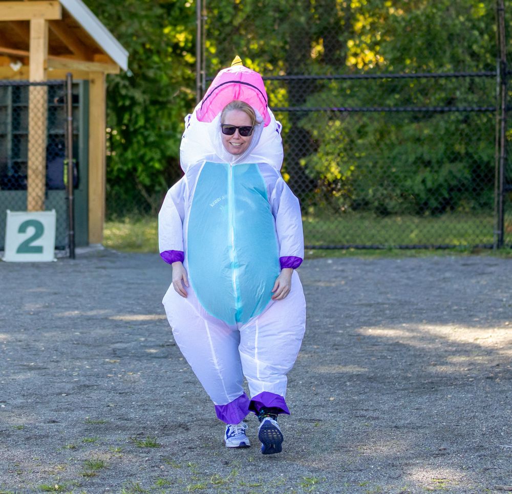 Person wearing inflatable unicorn costume walking on outdoor recreational field on sunny day.