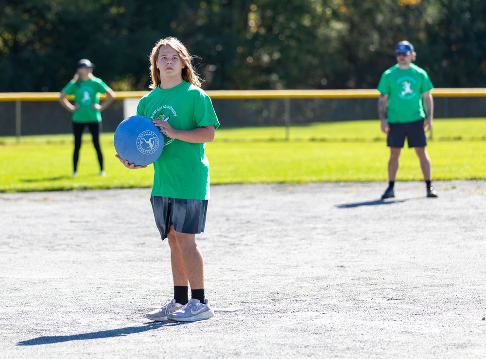 A young player in a green shirt holds a blue kickball on a sunny outdoor field during a game.