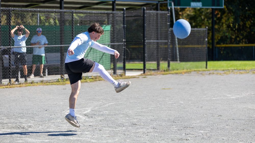 A young man kicks a blue balloon on an outdoor sports court on a sunny day.