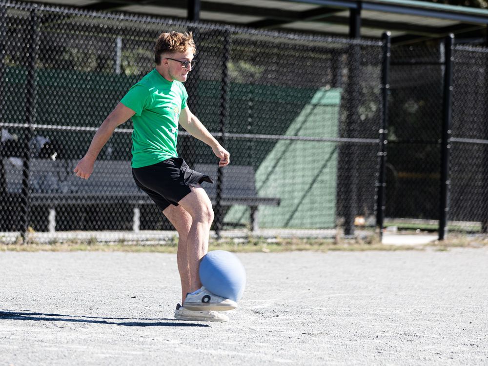 A child in a green shirt kicks a blue kickball on a sunny paved outdoor court during a game.