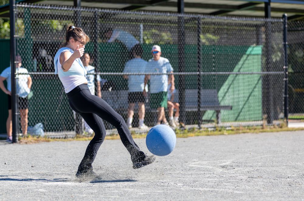A woman in a white shirt kicks a blue kickball on a sunny paved outdoor court during a game.