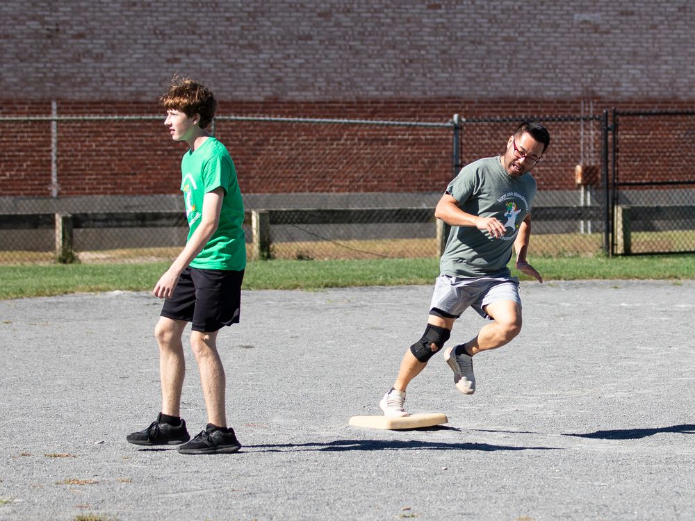 Two teenage boys play kickball on a paved outdoor court near a brick building.