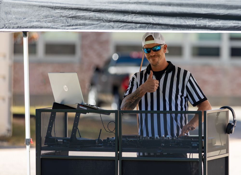 A DJ in a referee shirt gives thumbs up while mixing music on a laptop at an outdoor event.