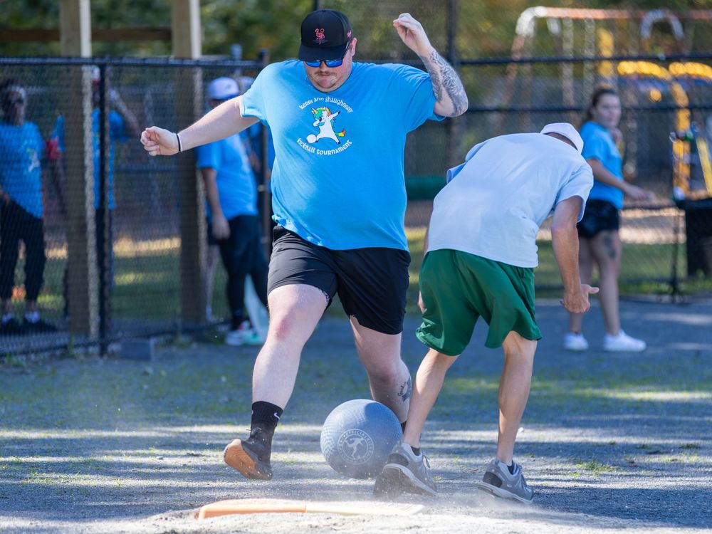 Two players compete for a gray kickball on a paved court during an outdoor kickball game.