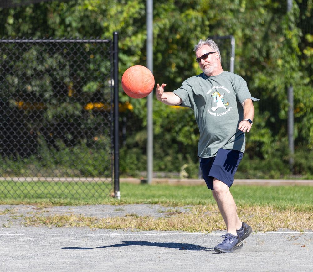 An older man in a gray shirt throws an orange ball on a paved outdoor court on a sunny day.