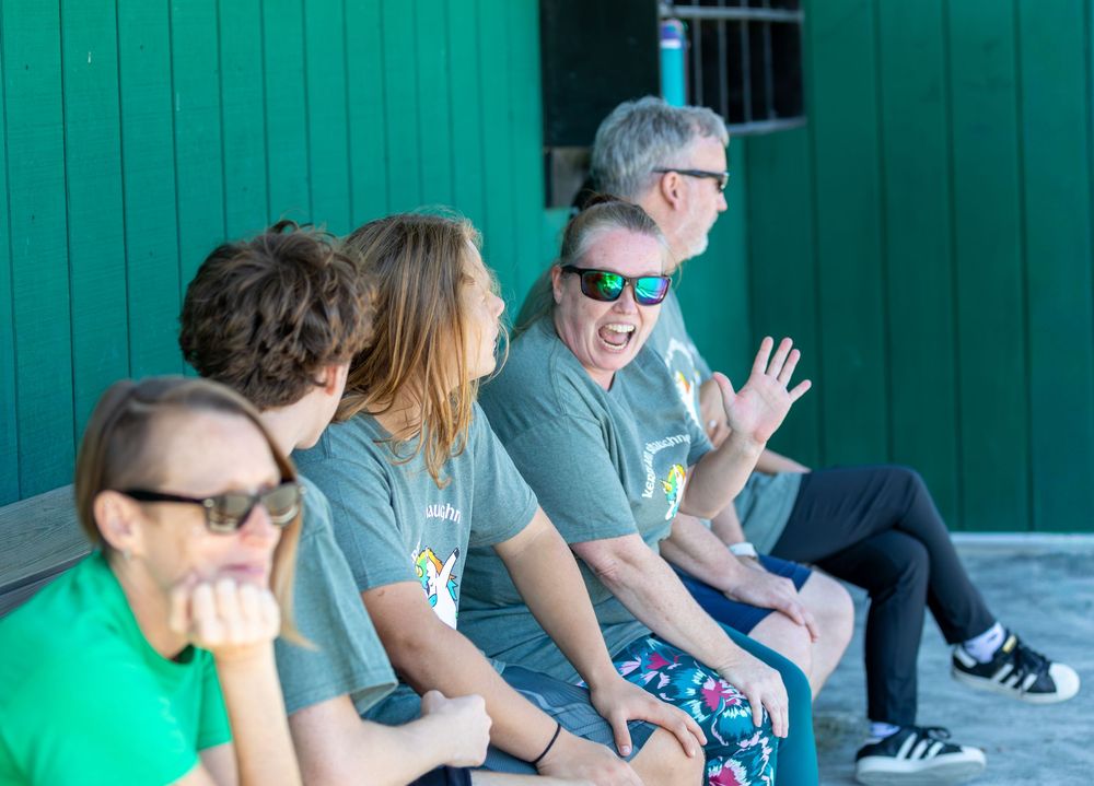 A group of adults in matching gray shirts laugh and chat while sitting on a bench outdoors.