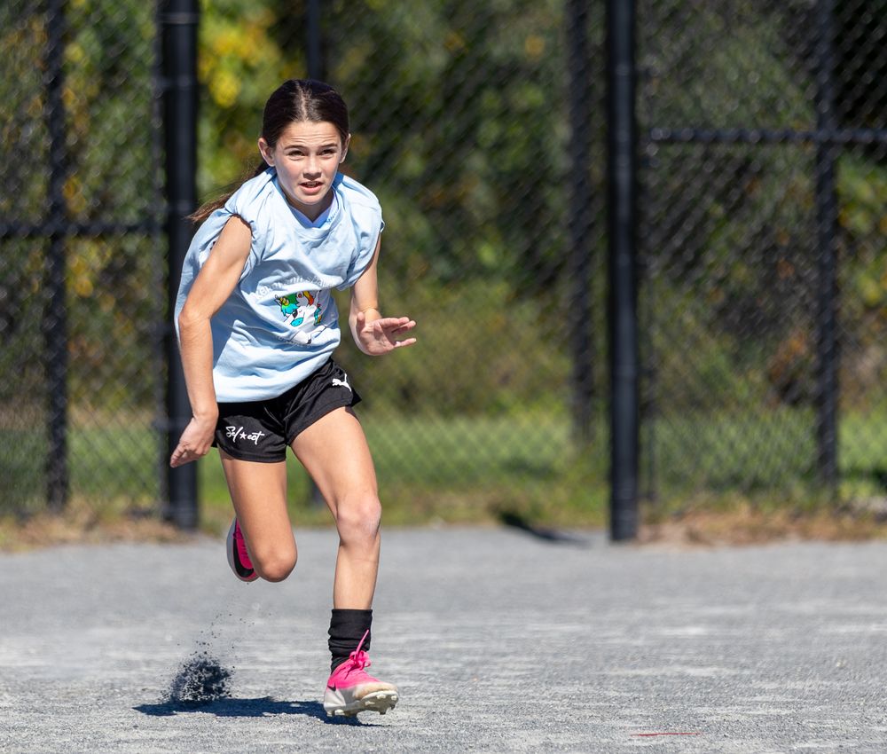 A girl in a light blue shirt sprints across a paved court wearing pink cleats during a game.