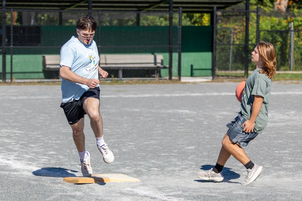Two young players run the bases on a sunny paved field during an outdoor kickball game.