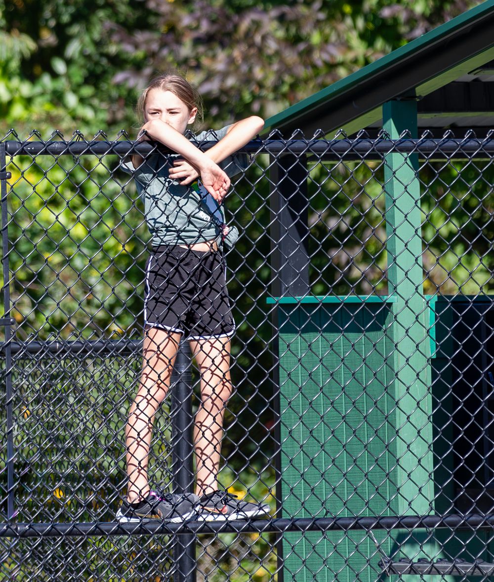 Young girl standing on chain-link fence outdoors shielding eyes from sun on bright day.
