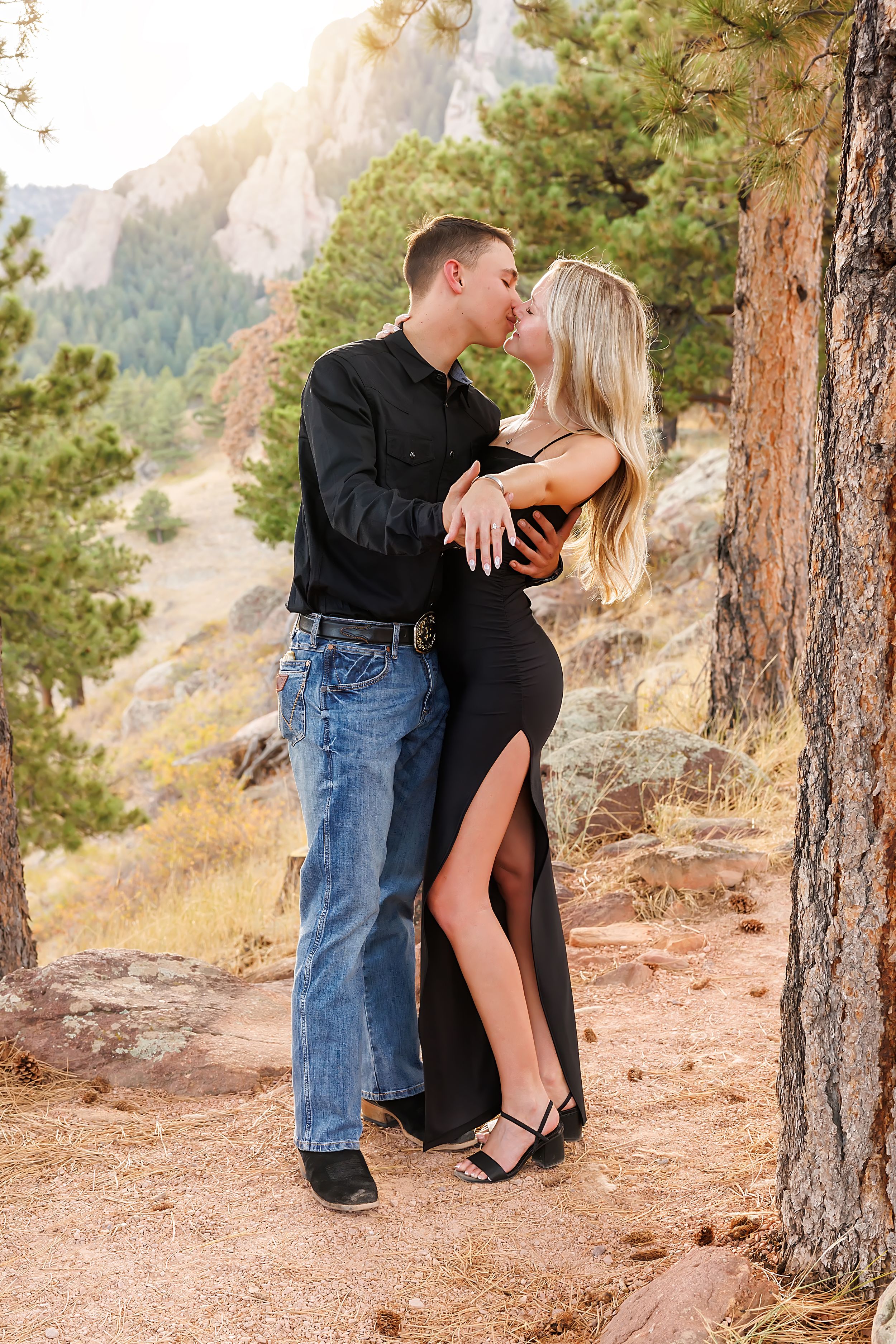 Couple kissing in a romantic outdoor engagement photo among pine trees with rocky mountain backdrop.