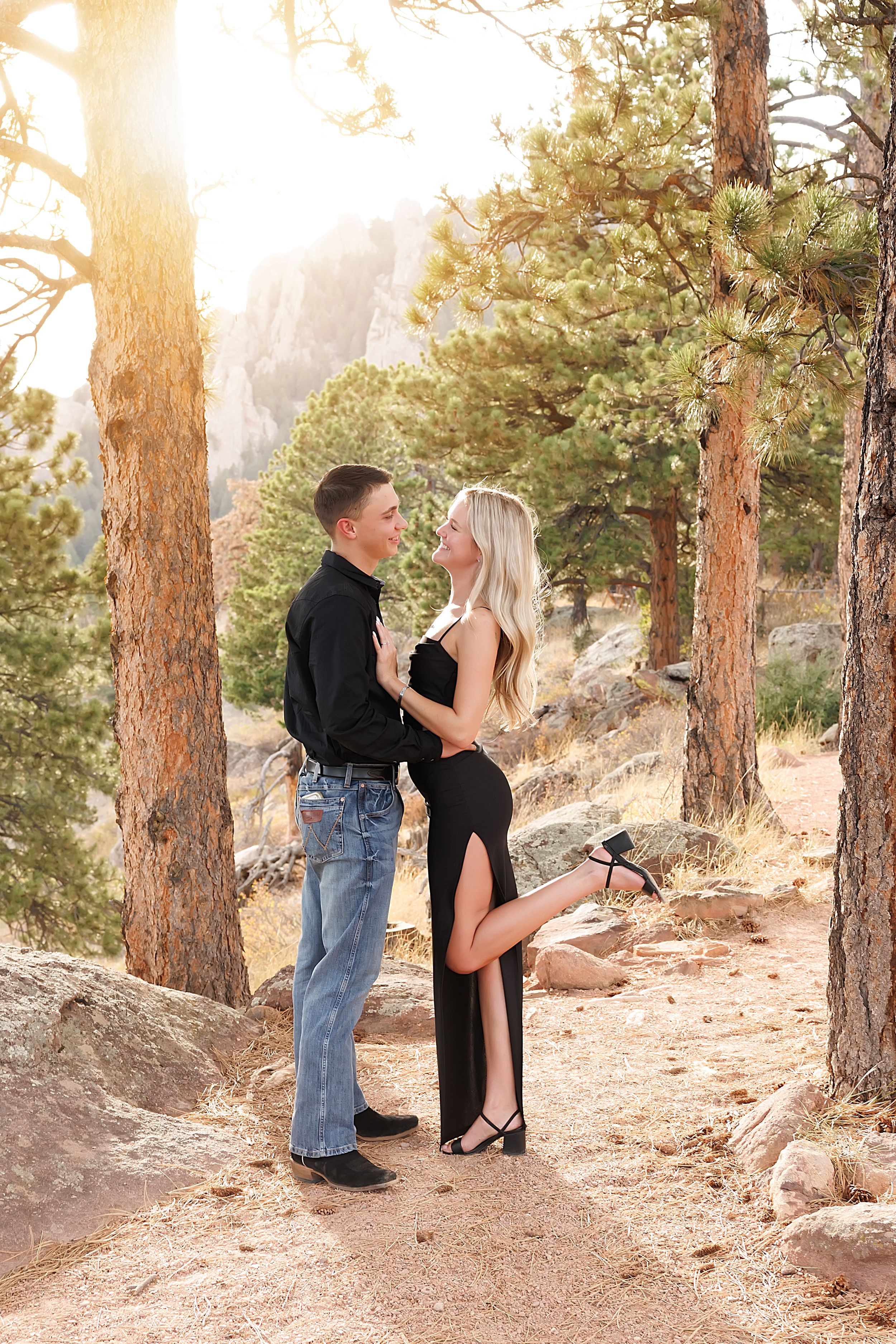 Couple in black outfits embracing among pine trees with golden sunlight in a rocky mountain forest setting.