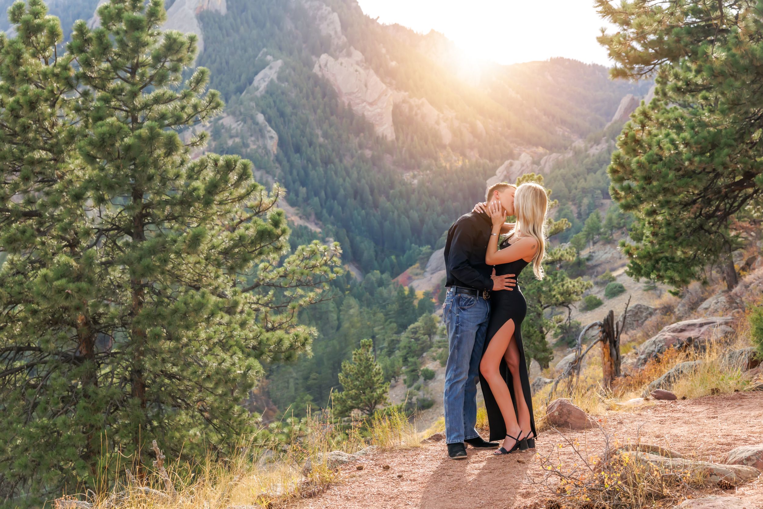 Couple embracing on a mountain trail surrounded by pine trees with sunlit rocky peaks in the background.