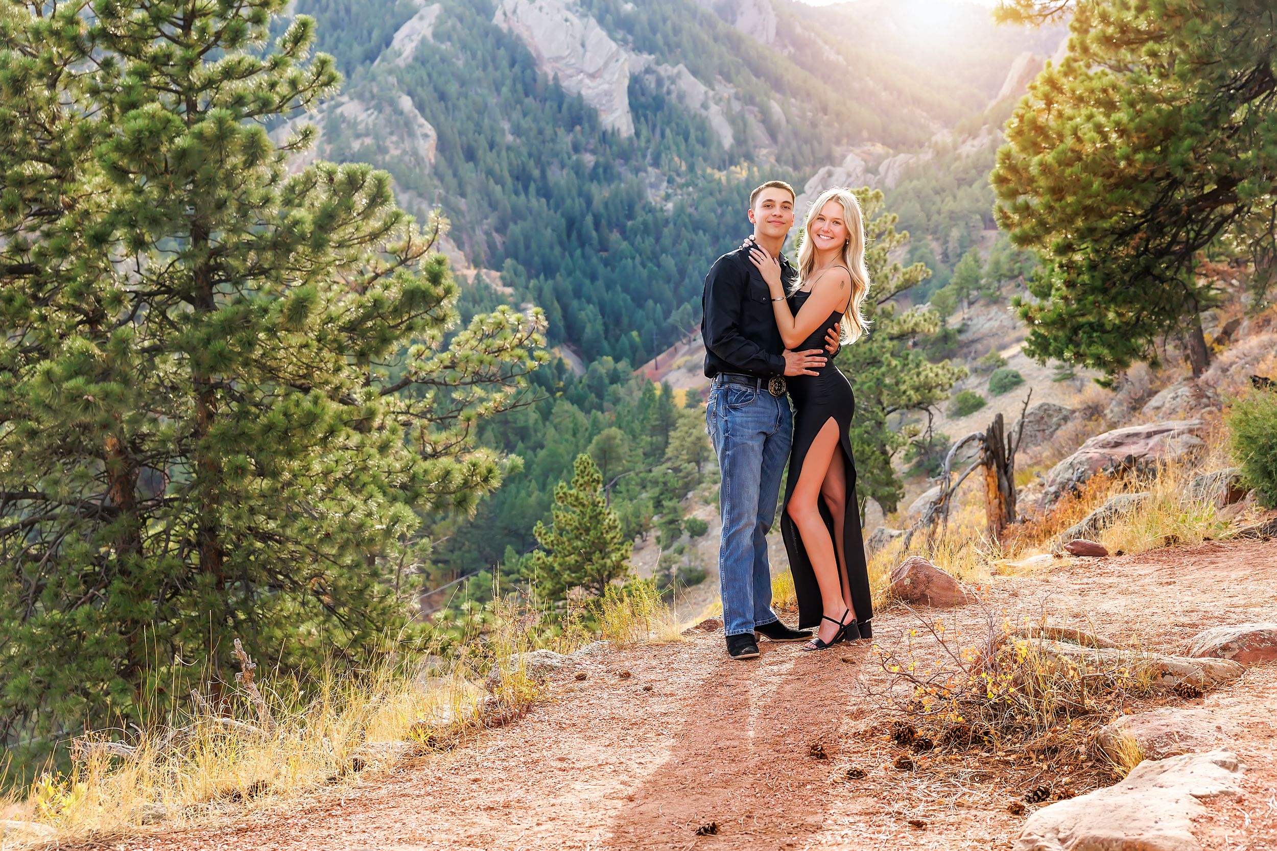 Couple embracing on a mountain trail surrounded by pine trees and rocky cliffs in a scenic outdoor engagement photo.