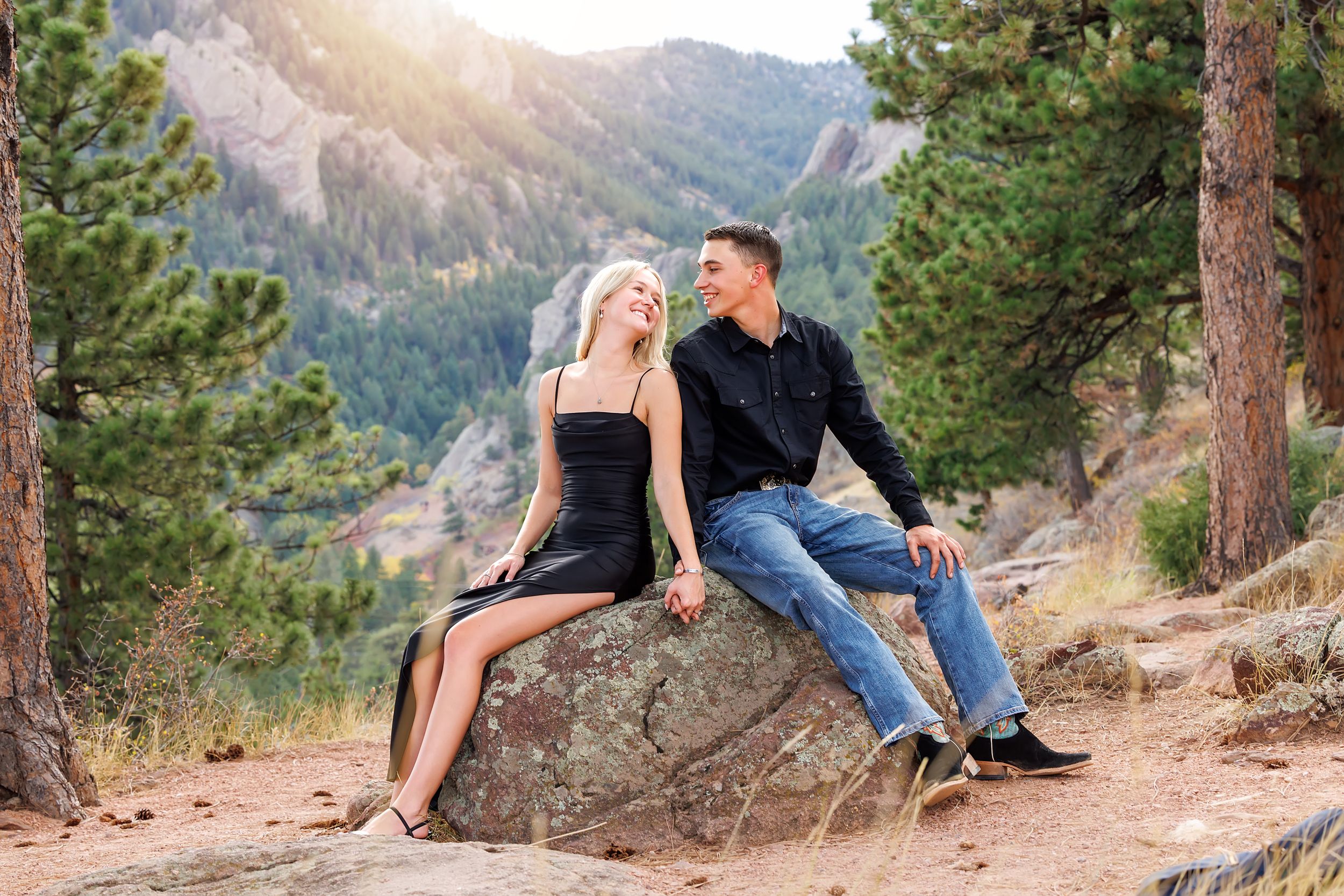 Couple sitting on a rock in a mountain forest, woman in black dress and man in jeans smiling together.