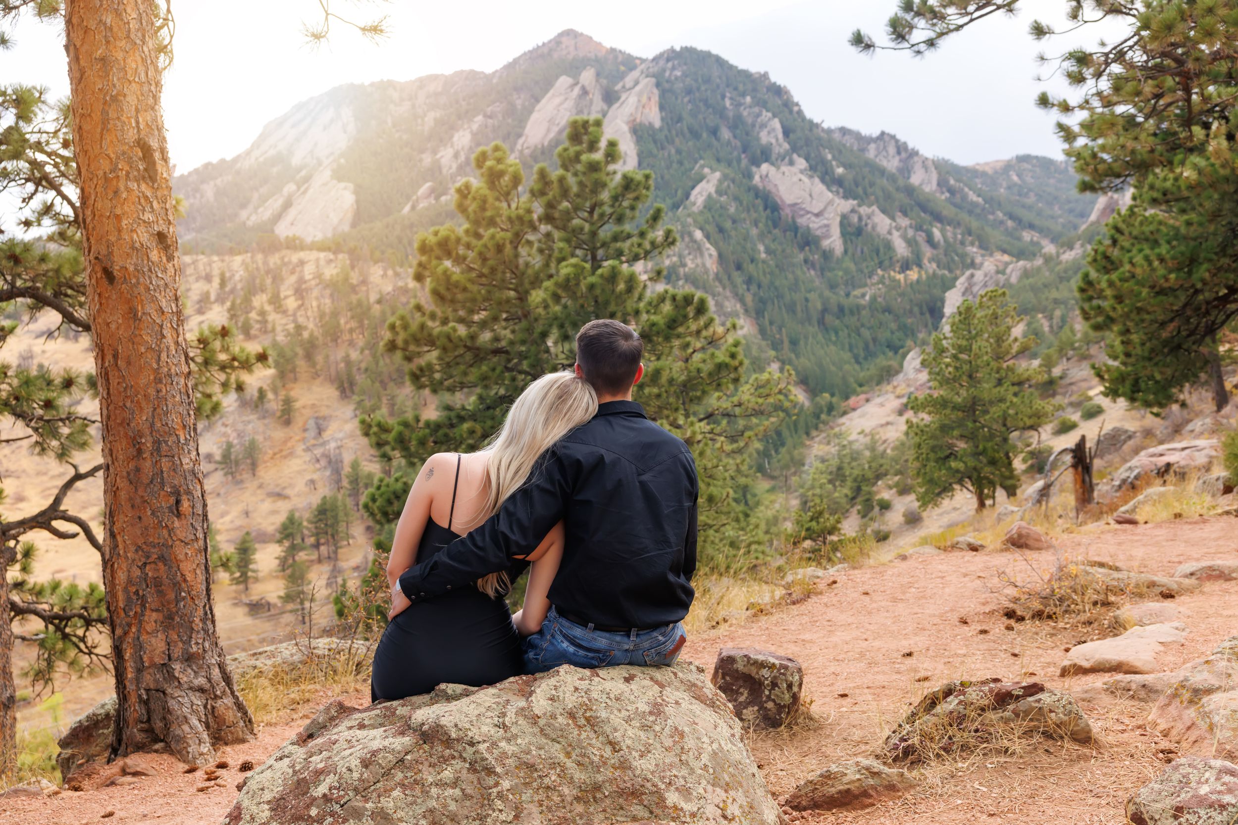Couple sitting on a boulder enjoying a scenic mountain view surrounded by pine trees and rocky peaks.
