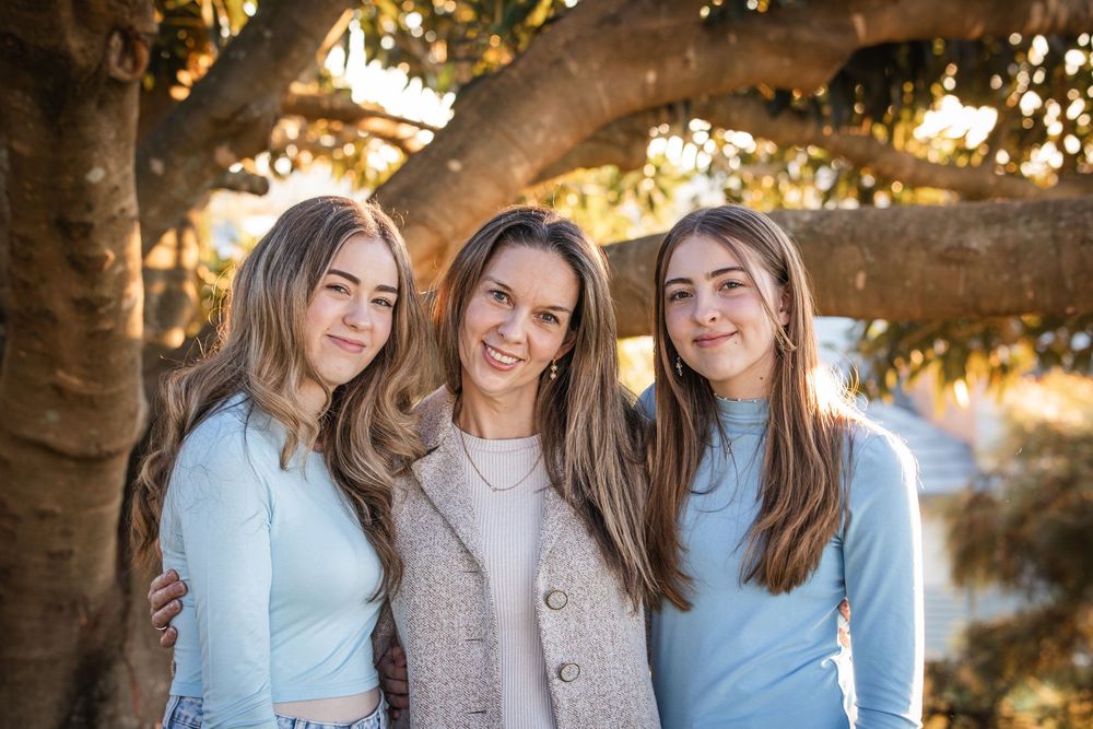 Three smiling young women with long hair pose arm in arm beneath a large tree branch in warm golden afternoon light in Beaumont Hills.