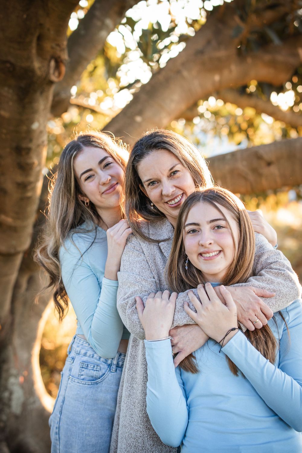 Three smiling women in light blue and soft neutral outfits embrace closely beneath a large tree in warm golden sunlight in Beaumont Hills.