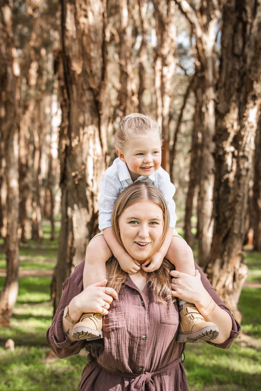 Smiling woman in a brown button-up dress holds a laughing toddler on her shoulders among tree trunks in a sunny forest in Beaumont Hills.