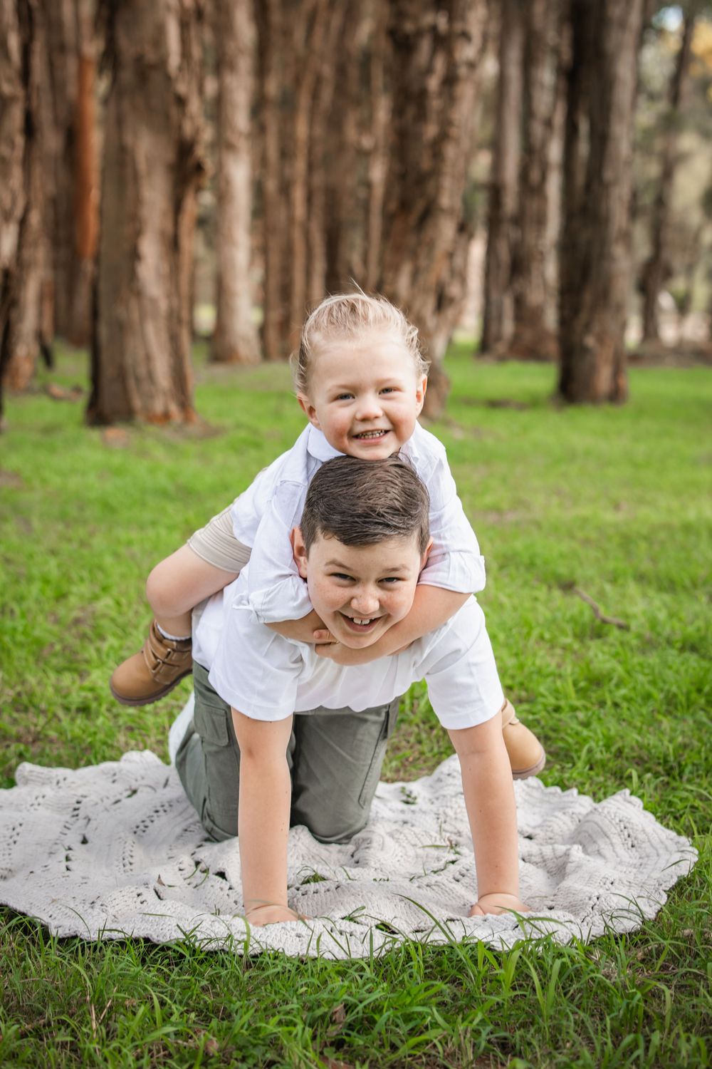 Two smiling children play on a lace blanket in a grassy park, one giving the other a piggyback ride among tall trees in Beaumont Hills.