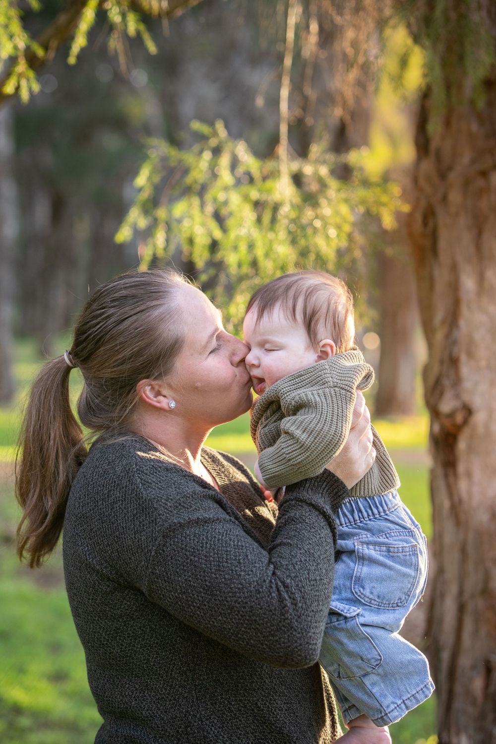 Mother with a ponytail kisses her baby's cheek while holding her beneath a large tree in warm golden afternoon light in Beaumont Hills.