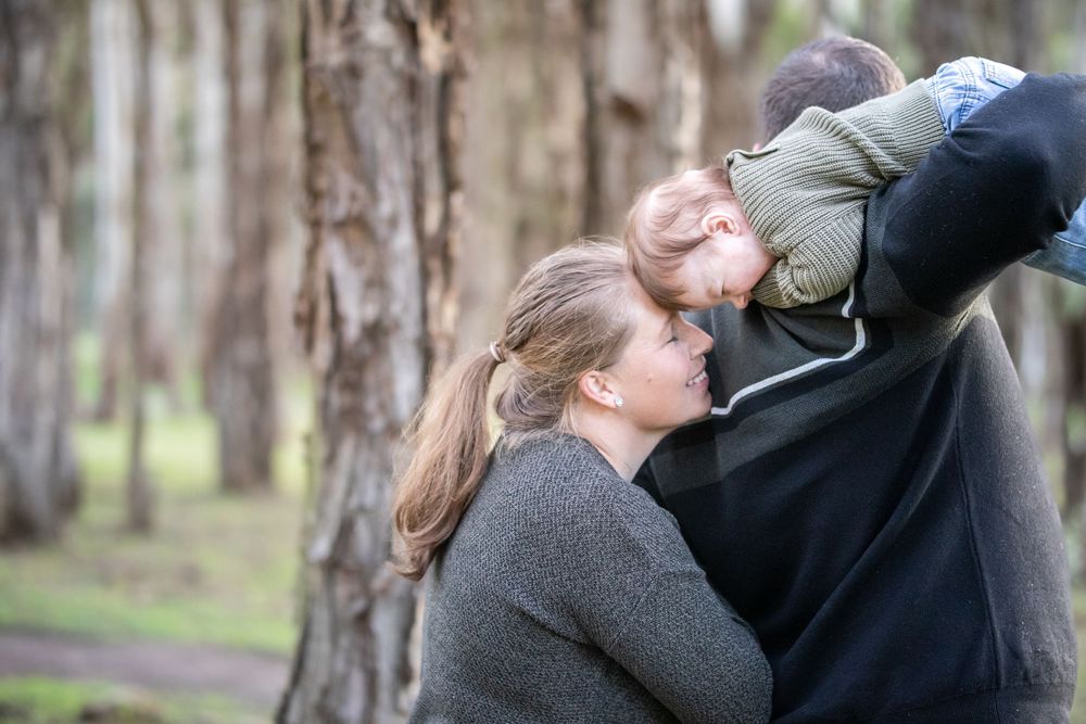 A woman with a ponytail leans her head against a man's chest as he holds a small child in a forest of tall trees in Beaumont Hills.