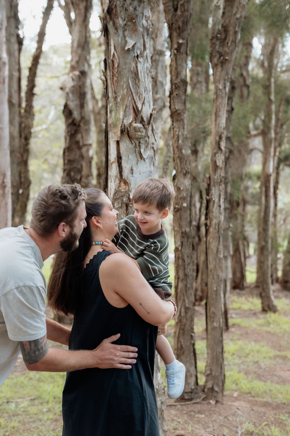 A smiling family of three stands among tall trees as a bearded man and a woman in a black dress hold their young son in Beaumont Hills.