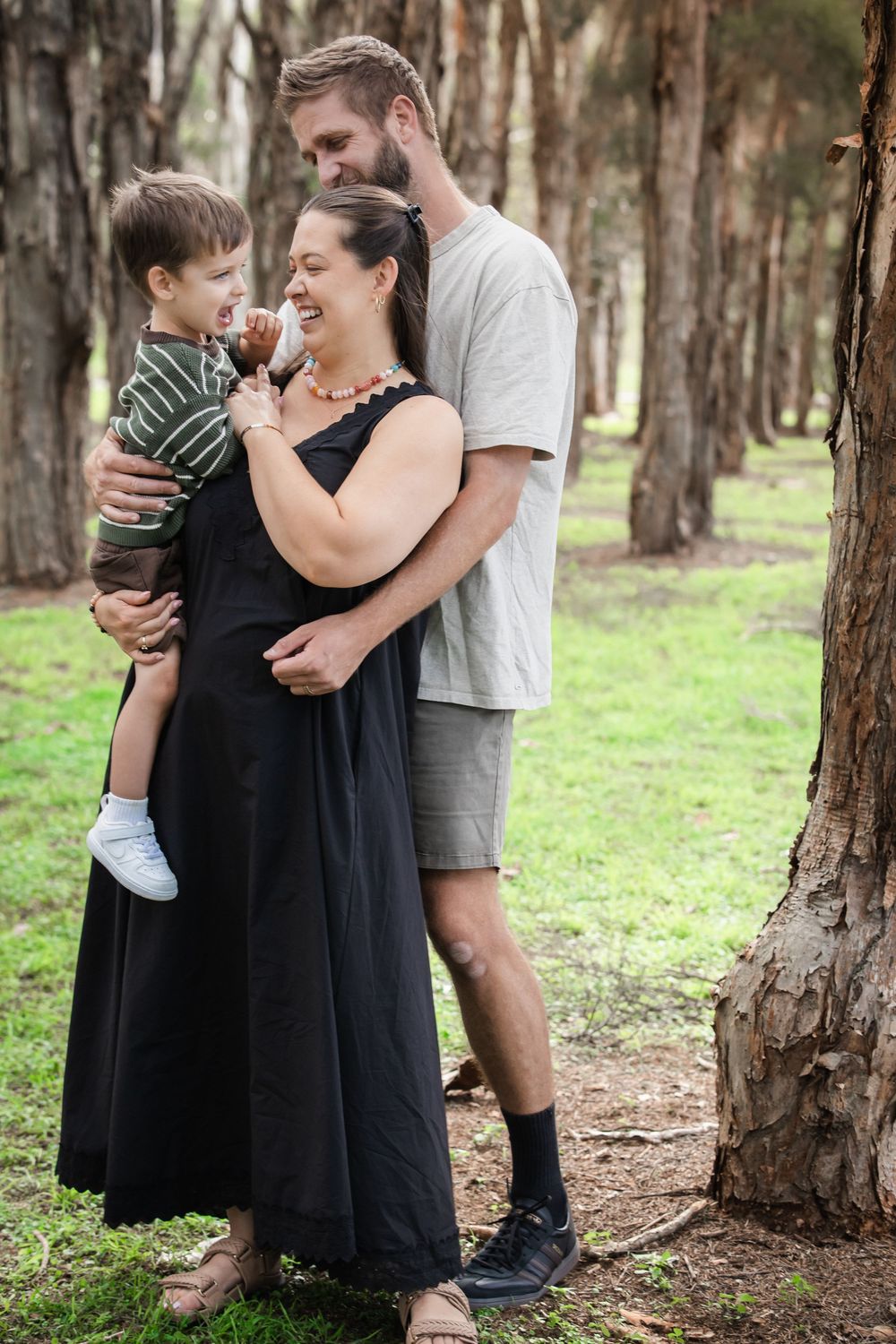 Smiling family of three embracing outdoors among tall trees, with a man hugging a woman in a black dress who holds a laughing toddler.