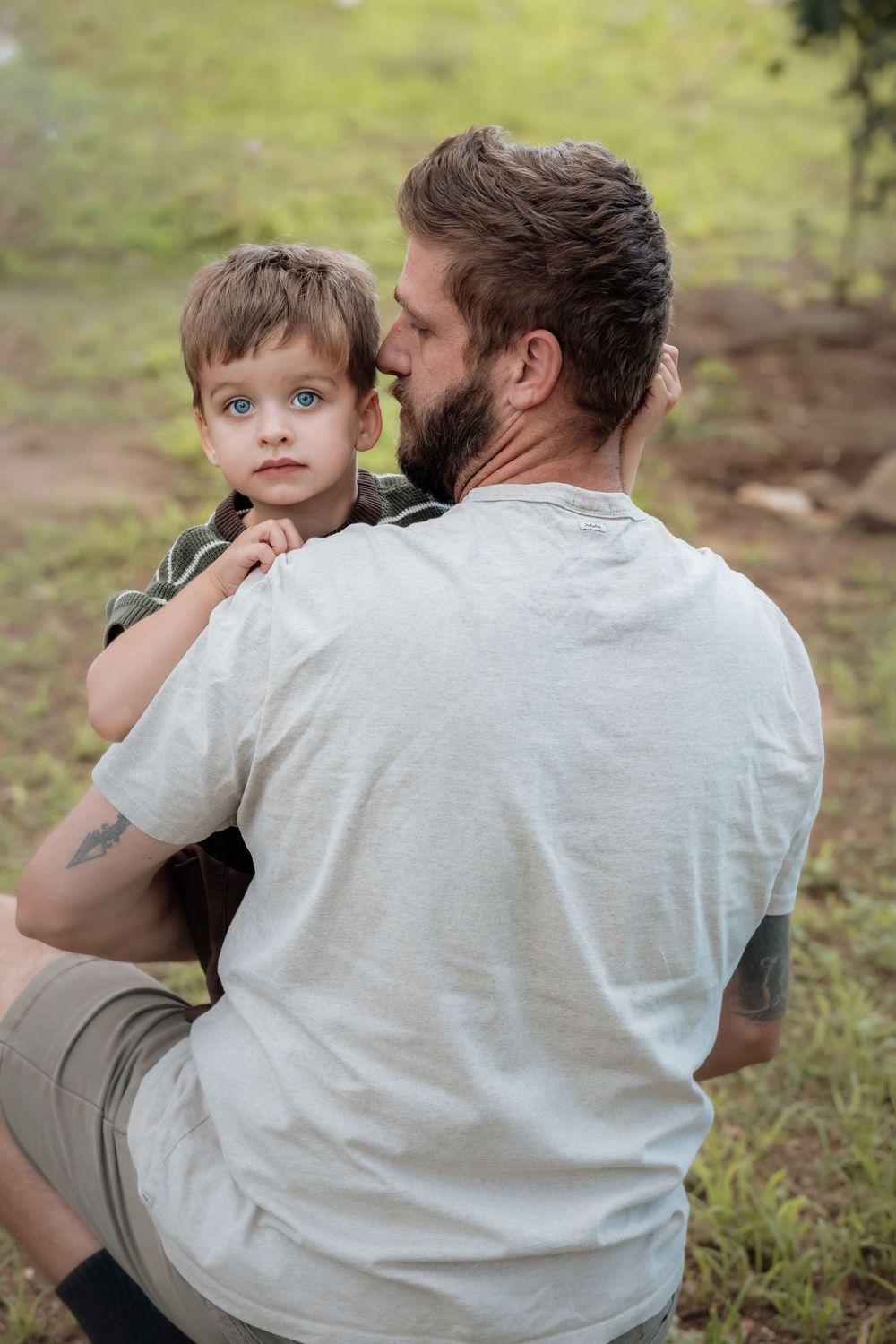 Bearded man in a gray t-shirt holds a blue-eyed young boy in his arms while standing on a grassy outdoor field in Beaumont Hills.