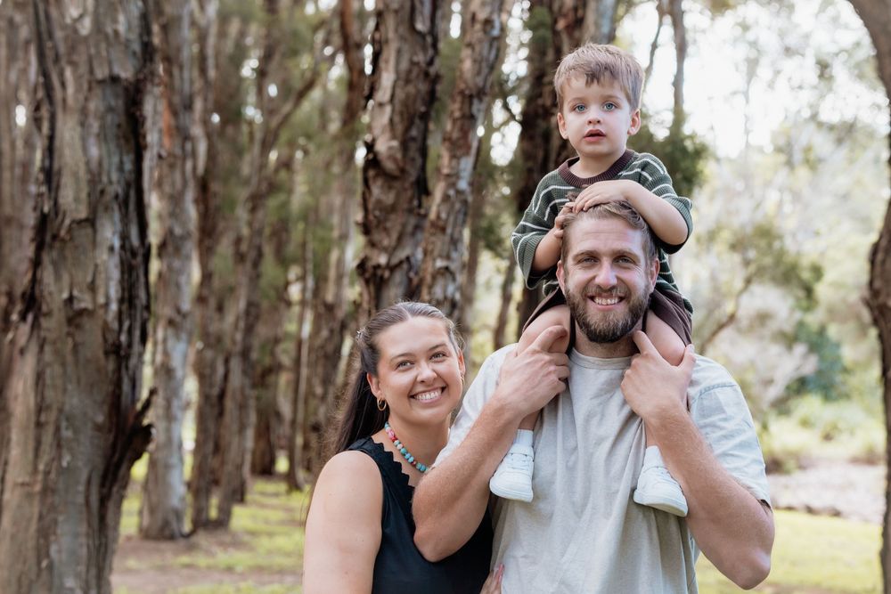 Smiling bearded man carrying a toddler on his shoulders beside a smiling woman among tall trees in a sunlit forest in Beaumont Hills.